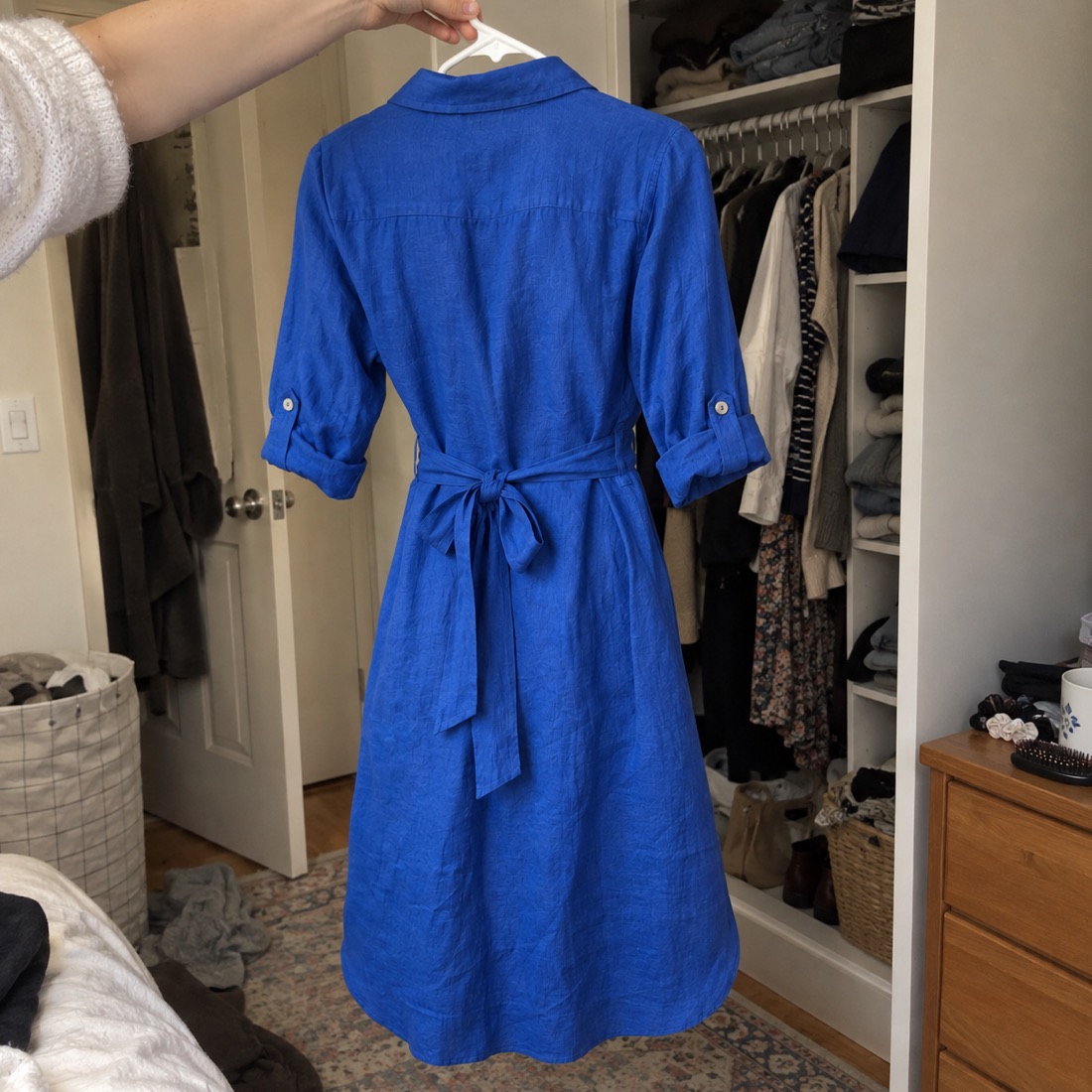 Phone snap of a blue linen shirt dress on a hanger in a bedroom